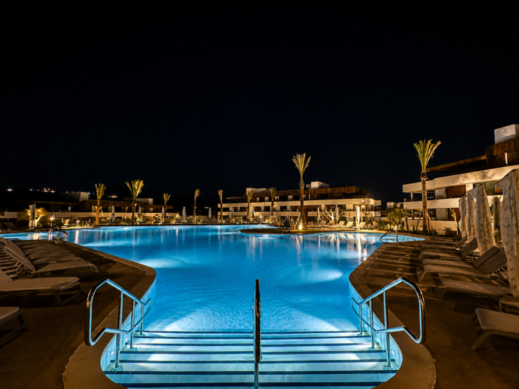 Piscina exterior de diseño en Village Verde, iluminada de noche, con tumbonas y palmeras.