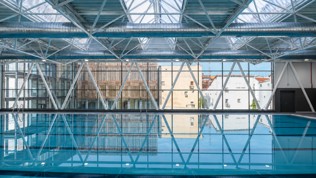 Piscina cubierta de competición con estructura metálica y reflejos en el agua.